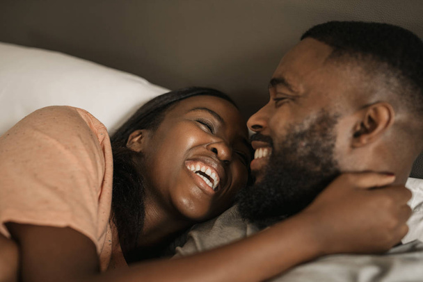 Affectionate young African American couple laughing and hugging each other while lying in bed together in the morning - Foto, imagen