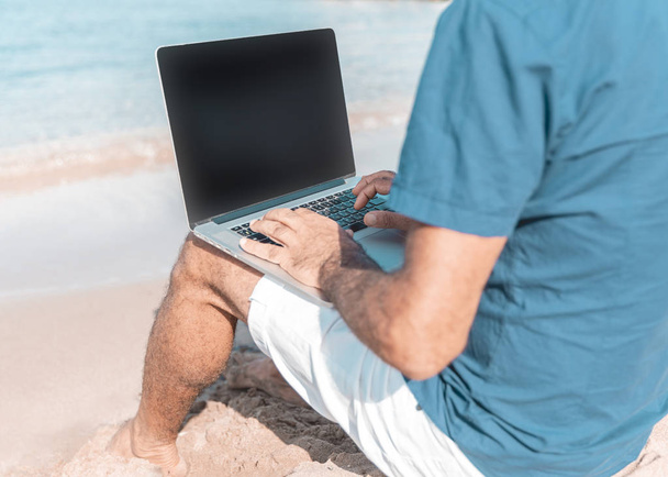 Hombre adulto de raza mediterránea usando portátil mientras está sentado en la playa. Primer plano de la pantalla del ordenador y las manos escribiendo en un teclado. Concepto de libertad y viaje
 - Foto, imagen