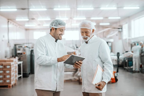 Young Caucasian supervisor showing results of food quality on tablet to his older colleague. Senior man holding folder with charts. Both are dressed in uniforms and having hairnets. Food plant. - Photo, Image