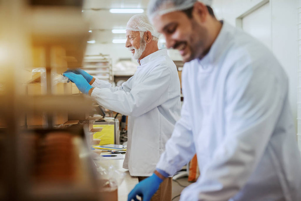 Funcionários caucasianos alegres vestidos com uniformes estéreis brancos embalando biscoitos em caixas enquanto estavam na fábrica de alimentos
. - Foto, Imagem