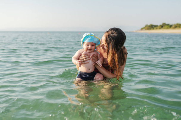 Совершила кавказскую брюнетку, обучая своего любящего 6-месячного сына плавать в море. Ребенок с шляпой на голове, наслаждаясь и улыбаясь
 - Фото, изображение
