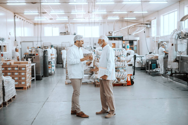 Two supervisors talking about quality of food. Younger one holding tablet while older one holding folder with charts. Both are dressed in sterile uniforms and having hairnets. Food plant interior. - Foto, Bild