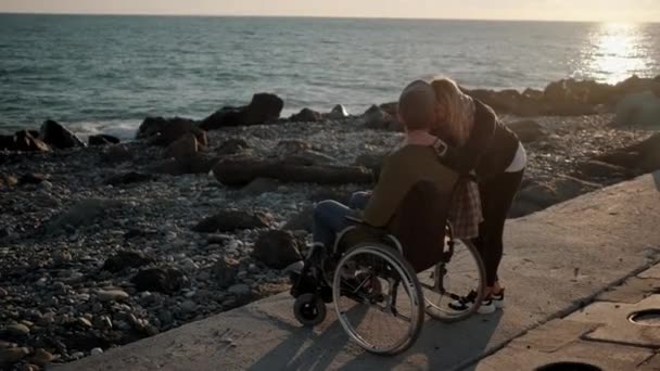 Chica sana está abrazando a su novio enfermo en silla de ruedas en muelle de mar
 - Metraje, vídeo