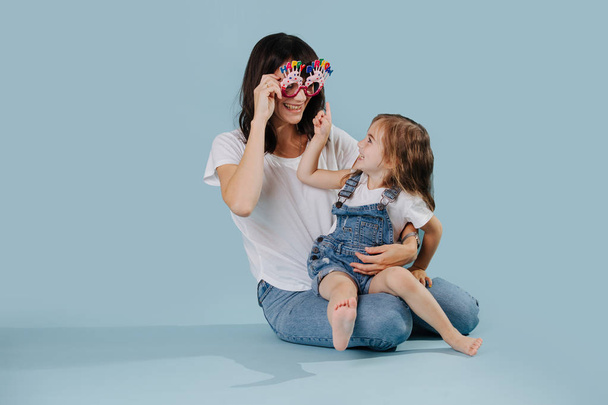 Mother and daughter playing with happy birthday glasses over blue background - Photo, Image