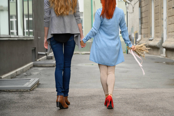 Stroll. Two girlfriends are walking along the street holding hands. The backs of two beautiful women on a walk with a bouquet of dried flowers. crumpled things - Foto, imagen