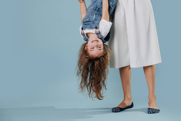 Matka trzyma jej śmiejąc córkę do góry nogami na niebieskim tle - Zdjęcie, obraz