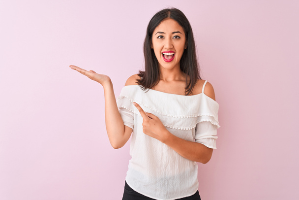 Beautiful chinese woman wearing white t-shirt standing over isolated pink background amazed and smiling to the camera while presenting with hand and pointing with finger. - Photo, Image