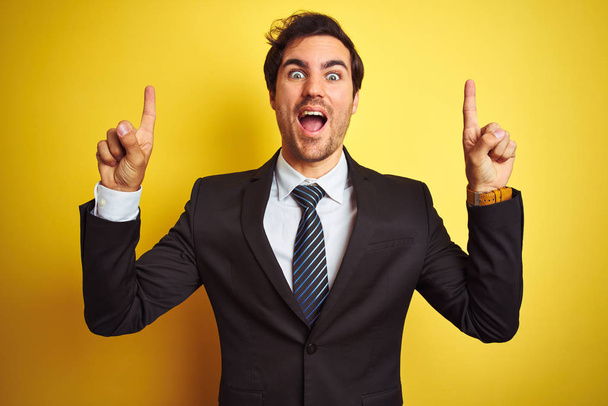 Young handsome businessman wearing suit and tie standing over isolated yellow background smiling amazed and surprised and pointing up with fingers and raised arms. - Foto, Bild