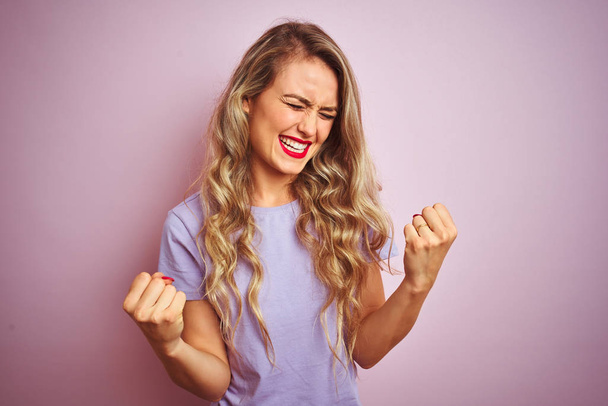 Young beautiful woman wearing purple t-shirt standing over pink isolated background very happy and excited doing winner gesture with arms raised, smiling and screaming for success. Celebration concept. - Zdjęcie, obraz