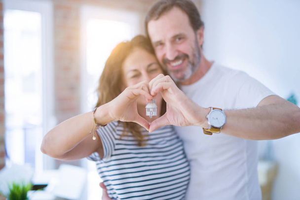 Edad media pareja romántica senior sosteniendo y mostrando las llaves de la casa, haciendo forma de corazón con las manos sonriendo feliz por mudarse a un nuevo hogar
 - Foto, Imagen