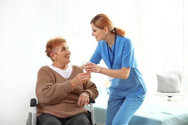Nurse giving glass of water to elderly woman in wheelchair indoors. Medical assistance - Foto, Imagem