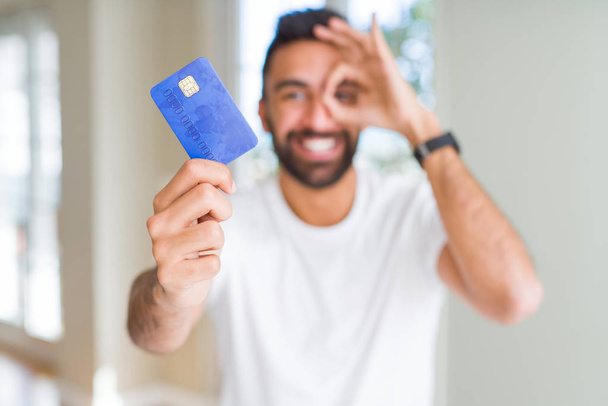 Bello ispanico uomo in possesso di carta di credito con volto felice sorridente facendo ok segno con mano sugli occhi guardando attraverso le dita
 - Foto, immagini