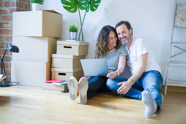 Średnim wieku starszy romantyczna para w miłości siedzi na podłodze w mieszkaniu z pudełka wokół i korzystania z komputera laptop uśmiechnięty szczęśliwy do przejścia do nowego domu - Zdjęcie, obraz