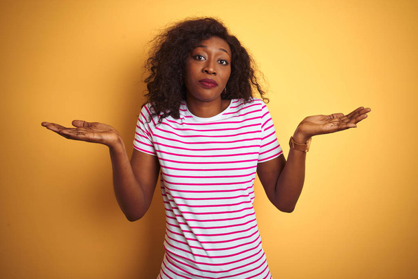 Młoda Afroamerykanka ubrana w koszulkę w paski na odizolowanym żółtym tle, nieświadoma i zdezorientowana, z podniesionymi rękami i rękami. Koncepcja wątpliwości. - Zdjęcie, obraz