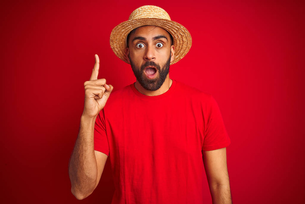 Jonge knappe indiaanse man draagt t-shirt en hoed over geïsoleerde rode achtergrond wijzend vinger omhoog met een succesvol idee. Opgewonden en gelukkig. Nummer één. - Foto, afbeelding