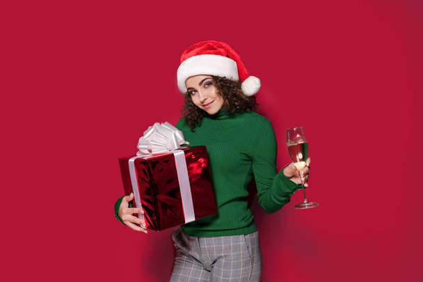 Beautiful fashion woman in santa claus hat, warm green Christmas sweater holding a glass of champagne and gift box on. - Valokuva, kuva
