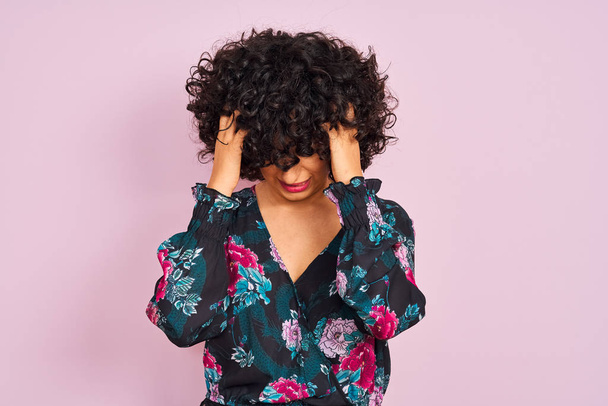 junge arabische Frau mit lockigem Haar trägt florales Kleid über isoliertem rosa Hintergrund und leidet unter Kopfschmerzen verzweifelt und gestresst, weil Schmerzen und Migräne. Hände auf den Kopf. - Foto, Bild
