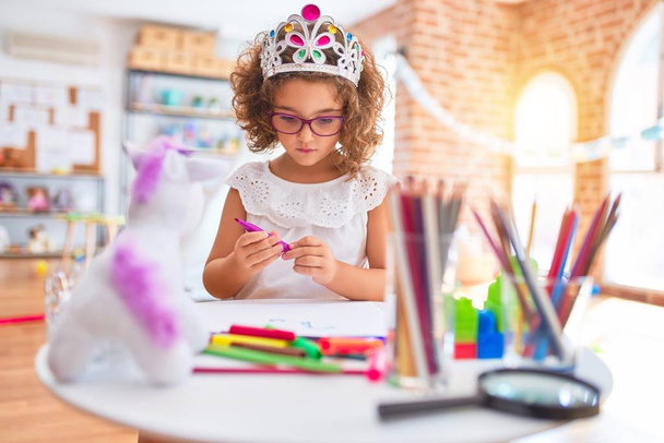 Piękny maluch nosi okulary i księżniczka korona siedzi rysunek przy użyciu papieru i markera pióra w przedszkolu - Zdjęcie, obraz