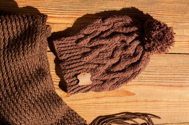 Brown knitted hat and scarf on a wooden background with natural light shadow. - Photo, Image