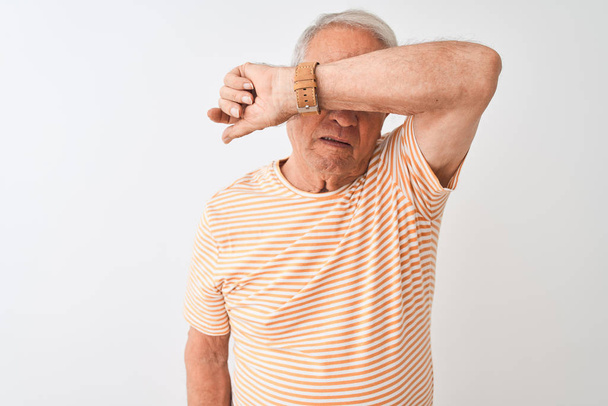 Ein älterer grauhaariger Mann mit gestreiftem T-Shirt steht vor isoliertem weißen Hintergrund und bedeckt die Augen mit einem Arm, sieht ernst und traurig aus. Versteck- und Ablehnungskonzept - Foto, Bild