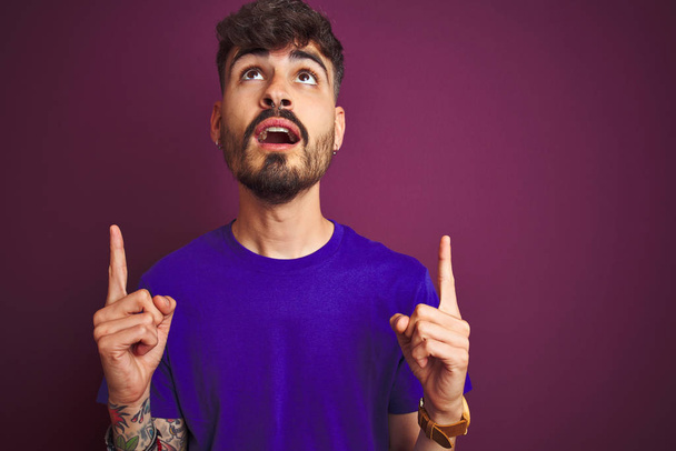 Молодой человек с татуировкой в футболке, стоящий на изолированном фиолетовом фоне удивлен и удивлен, глядя вверх и указывая пальцами и поднятыми руками
. - Фото, изображение
