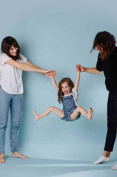 Two adults lifting little girl by hands for her amusement over blue background - Foto, afbeelding