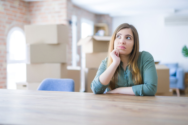 Young woman sitting on the table with cardboard boxes behind her moving to new home with hand on chin thinking about question, pensive expression. Smiling with thoughtful face. Doubt concept. - Photo, image