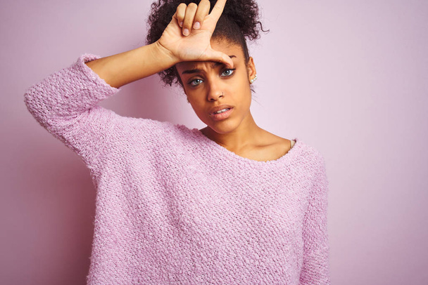 Молода афроамериканка у зимовому светрі, що стоїть над рожевим фоном, сміється з людей з пальцями на лобі, глузує і ображає.. - Фото, зображення