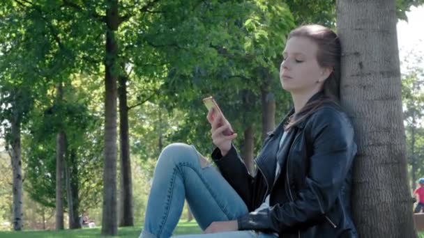 Young beautiful girl in street clothes in a park. Holding a smartphone in his hands, sitting on the grass under a tree. Reads boring message and photos, Yawns heavily. Not enough sleep, wants to sleep - 映像、動画