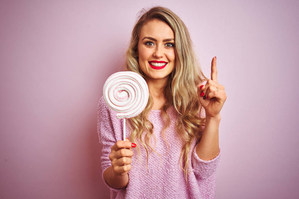Jovem mulher bonita comendo doces doces sobre rosa isolado fundo surpreso com uma ideia ou pergunta apontando dedo com o rosto feliz, número um
 - Foto, Imagem