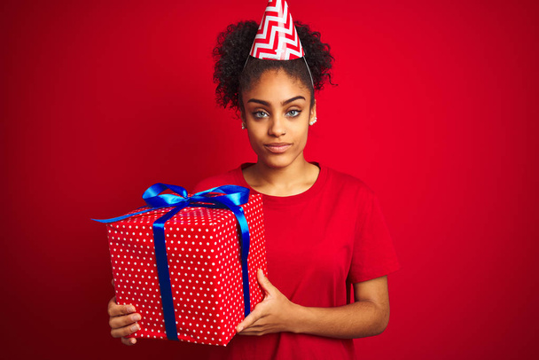 African american woman wearing funny hat holding birthday gift over isolated red background with a confident expression on smart face thinking serious - Fotografie, Obrázek