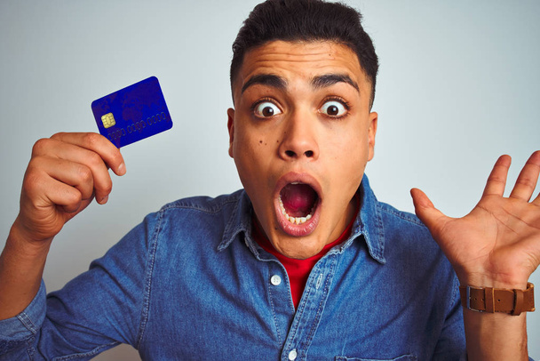 Jeune homme client brésilien tenant la carte de crédit debout sur fond blanc isolé très heureux et excité, expression gagnante célébrant la victoire en criant avec un grand sourire et les mains levées
 - Photo, image