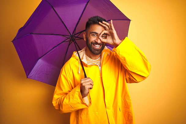 Young indian man wearing raincoat and purple umbrella over isolated yellow background with happy face smiling doing ok sign with hand on eye looking through fingers - Фото, изображение