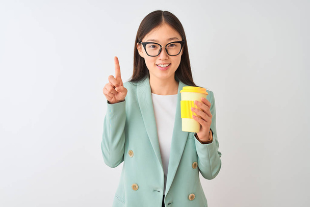 junge Chinesin trägt Brille Kaffee trinken über isolierten weißen Hintergrund überrascht mit einer Idee oder Frage Zeigefinger mit glücklichem Gesicht, Nummer eins - Foto, Bild