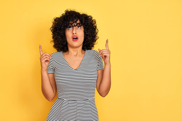 Young arab woman with curly hair wearing striped dress over isolated yellow background amazed and surprised looking up and pointing with fingers and raised arms. - Valokuva, kuva
