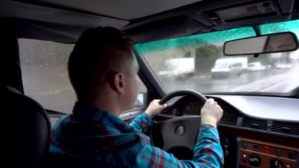 Mladý muž řídí auto. Deštivé počasí. Pohled na muže zezadu ze zadního sedadla - Záběry, video