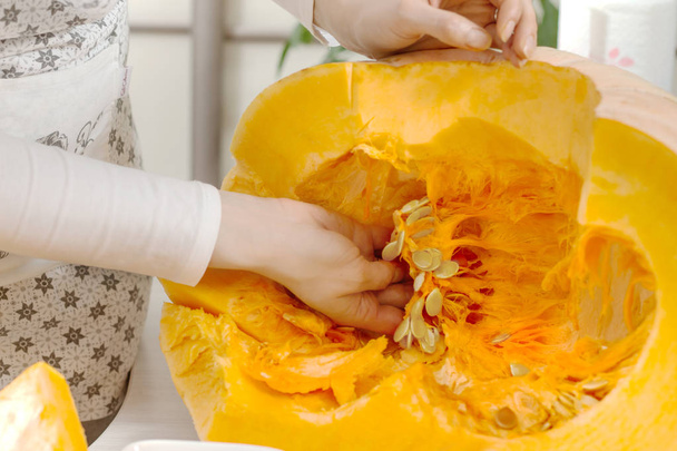Woman's hand chooses the pulp from the center of a large fresh pumpkin with seeds. A woman wearing a kitchen apron. A large pumpkin cut in half lies on the kitchen counter. - Photo, Image