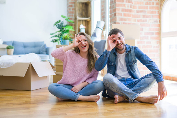 Young beautiful couple moving to a new house sitting on the floor doing ok gesture with hand smiling, eye looking through fingers with happy face. - Fotoğraf, Görsel