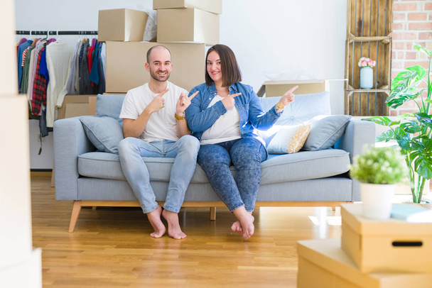 Jovem casal sentado no sofá arround caixas de papelão movendo-se para uma nova casa sorrindo e olhando para a câmera apontando com duas mãos e dedos para o lado
. - Foto, Imagem