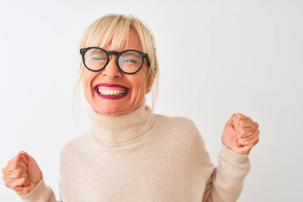 孤立した白い背景の上に亀甲のセーターと眼鏡を身に着けている中年の女性は非常に幸せと腕を上げて勝者のジェスチャーを行う興奮,笑顔と成功のために叫んで.お祝いのコンセプト. - 写真・画像