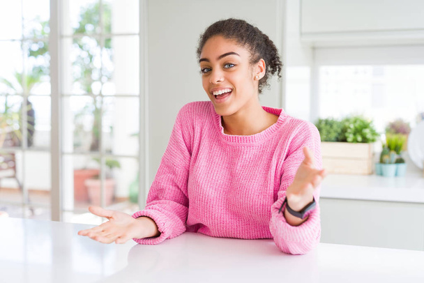 Hermosa mujer afroamericana con cabello afro vistiendo suéter rosa casual sonriendo alegre con los brazos abiertos como bienvenida amistosa, saludos positivos y confiados
 - Foto, imagen