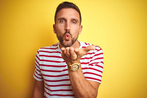 Young handsome man wearing casual red striped t-shirt over yellow isolated background looking at the camera blowing a kiss with hand on air being lovely and sexy. Love expression. - Foto, Imagen