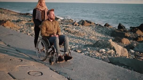 Young woman is nursing sick disabled man, rolling stroller on sea shore - Footage, Video