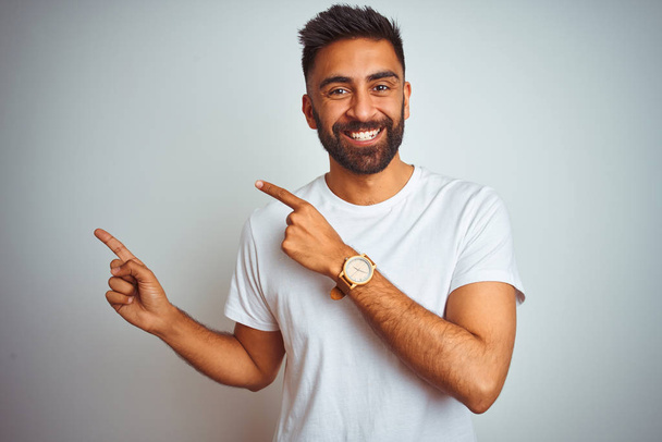 Молодий індійський чоловік у футболці стоїть над ізольованим білим тлом, посміхаючись і дивлячись на камеру, спрямовану двома руками і пальцями на бік
. - Фото, зображення