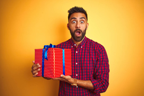 Arap Hintli İspanyol adamın elinde doğum günü hediyesi, izole edilmiş sarı arka planda duruyor. Şaşırmış bir yüzle korkmuş ve korku ifadesinden heyecanlanmış. - Fotoğraf, Görsel