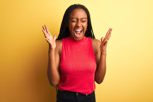 Африканська американка, одягнена в червону випадкову футболку, стоїть над ізольованим жовтим фоном, який прославляє божевілля і божевілля за успіх з піднятими руками і закритими очима, що кричать захоплено. Концепція переможця - Фото, зображення