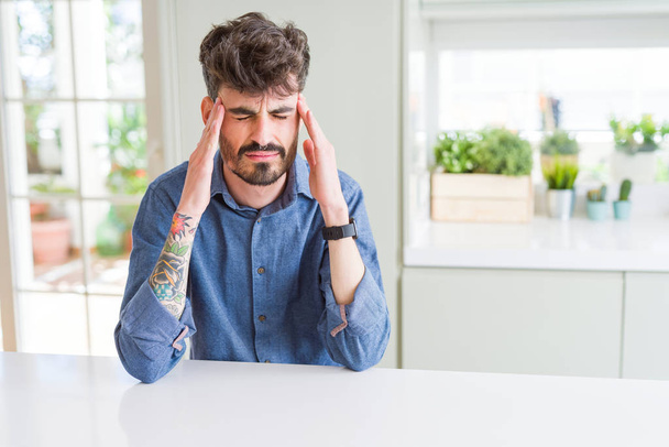 Jongeman draagt casual shirt zittend op witte tafel met de hand op het hoofd voor pijn in het hoofd omdat stress. Lijdende migraine. - Foto, afbeelding
