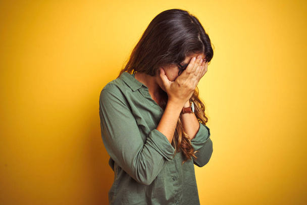 Young beautiful woman wearing green shirt and glasses over yelllow isolated background with sad expression covering face with hands while crying. Depression concept. - 写真・画像