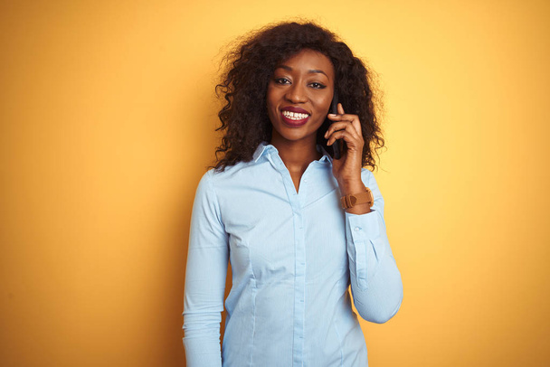 Genç Afro-Amerikan kadın akıllı telefondan konuşuyor. İzole edilmiş sarı arka plan üzerinde. Mutlu bir yüzü var ve gülümsüyor. - Fotoğraf, Görsel