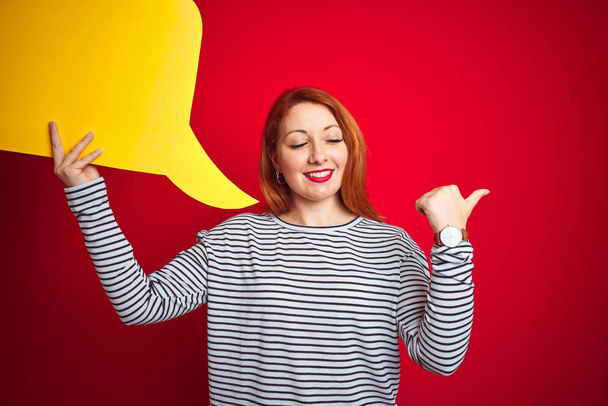 Fiatal gyönyörű vörös hajú nő kezében sárga beszéd buborék felett piros elszigetelt háttér mutató és mutatja hüvelykujjával felfelé az oldalon boldog arc mosolygós - Fotó, kép